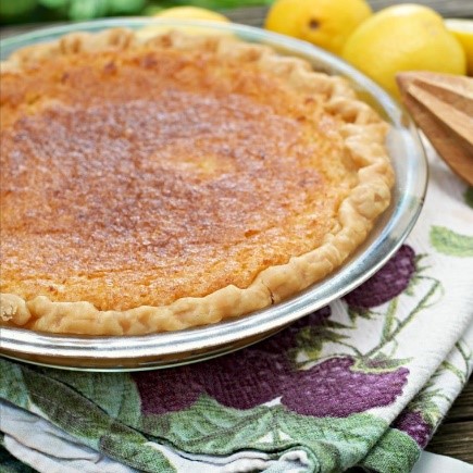 Fresh baked lemon chess pie with golden brown crust in a glass pie pan surrounded on one edge with fresh lemons